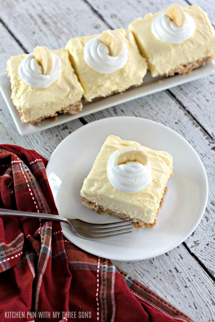 A top view of a square of banana cream cheesecake next to a dish towel, with three more squares in the background.